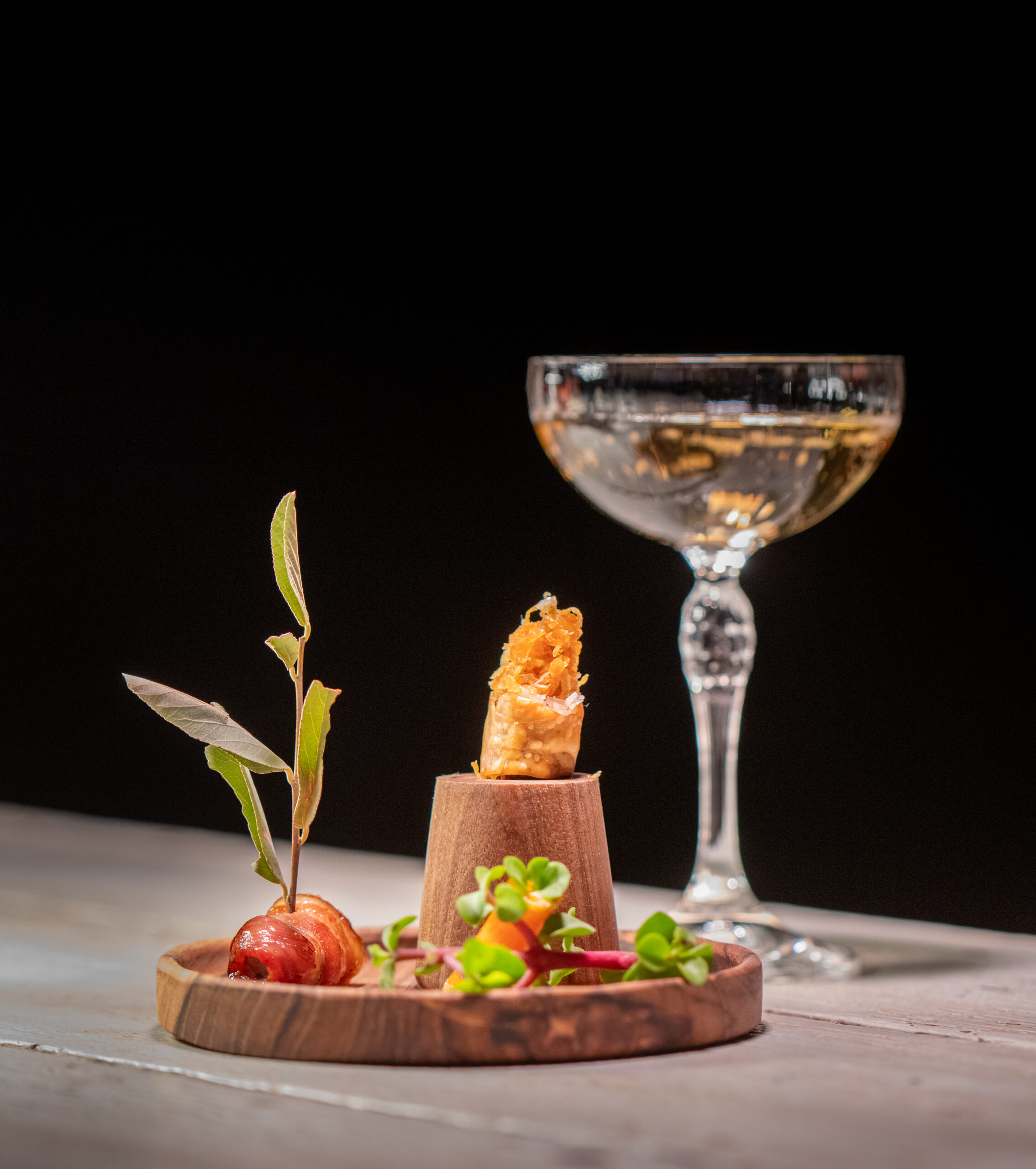 A gourmet dish on a wooden plate with a green garnish, beside a filled champagne coupe glass on a wooden surface against a black background.