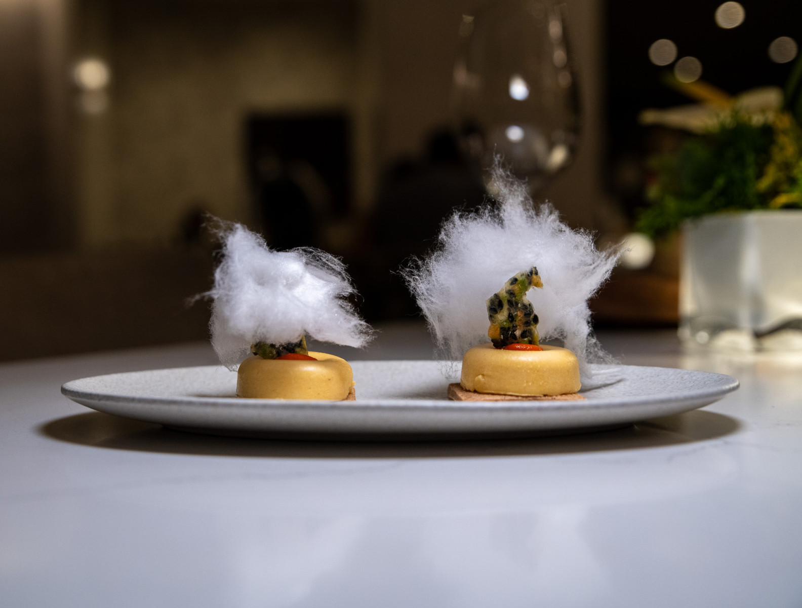 Two small round desserts topped with cotton candy and fruit are displayed on a white plate on a table in a dimly lit setting.
