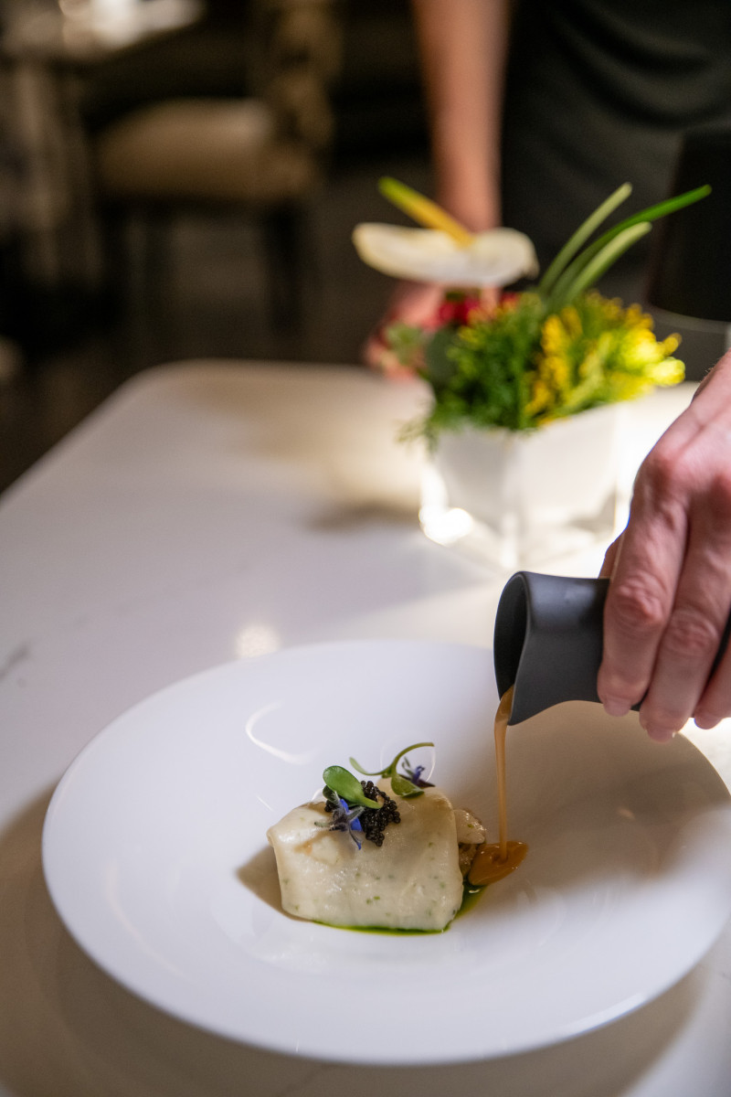 A hand pours sauce from a small pitcher onto a plated dish garnished with microgreens and edible flowers, with a floral arrangement in the background.