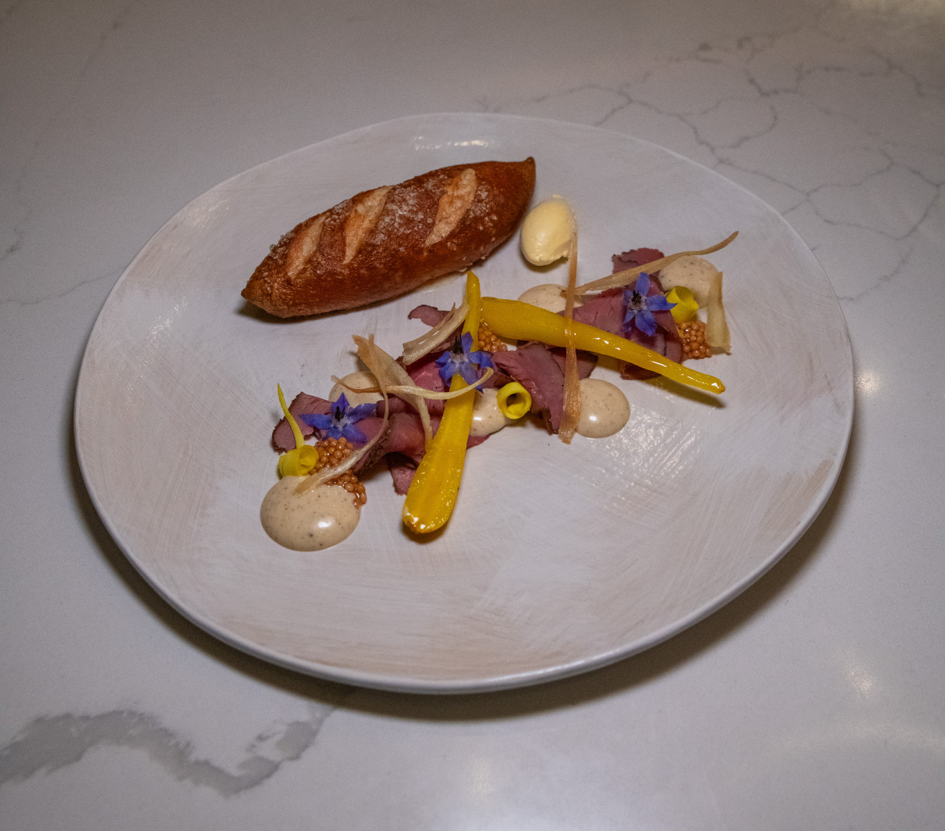 A plated dish with a small baguette, a dollop of butter, yellow carrots, thinly sliced meat, sauce, mustard seeds, and edible blue flowers on a white plate.