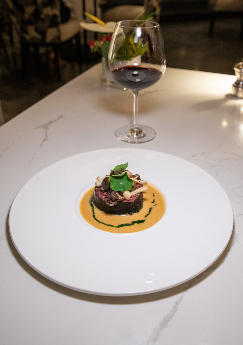 A plated gourmet dish with sauce and garnish is served on a white plate, accompanied by a glass of red wine on a marble table.